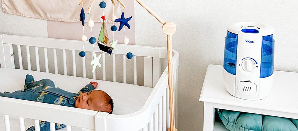 An infant is lying in a crib next to a Vicks humidifier for a baby