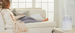 A woman sitting on a couch reading on a tablet, while a Vicks humidifier humidifies the room